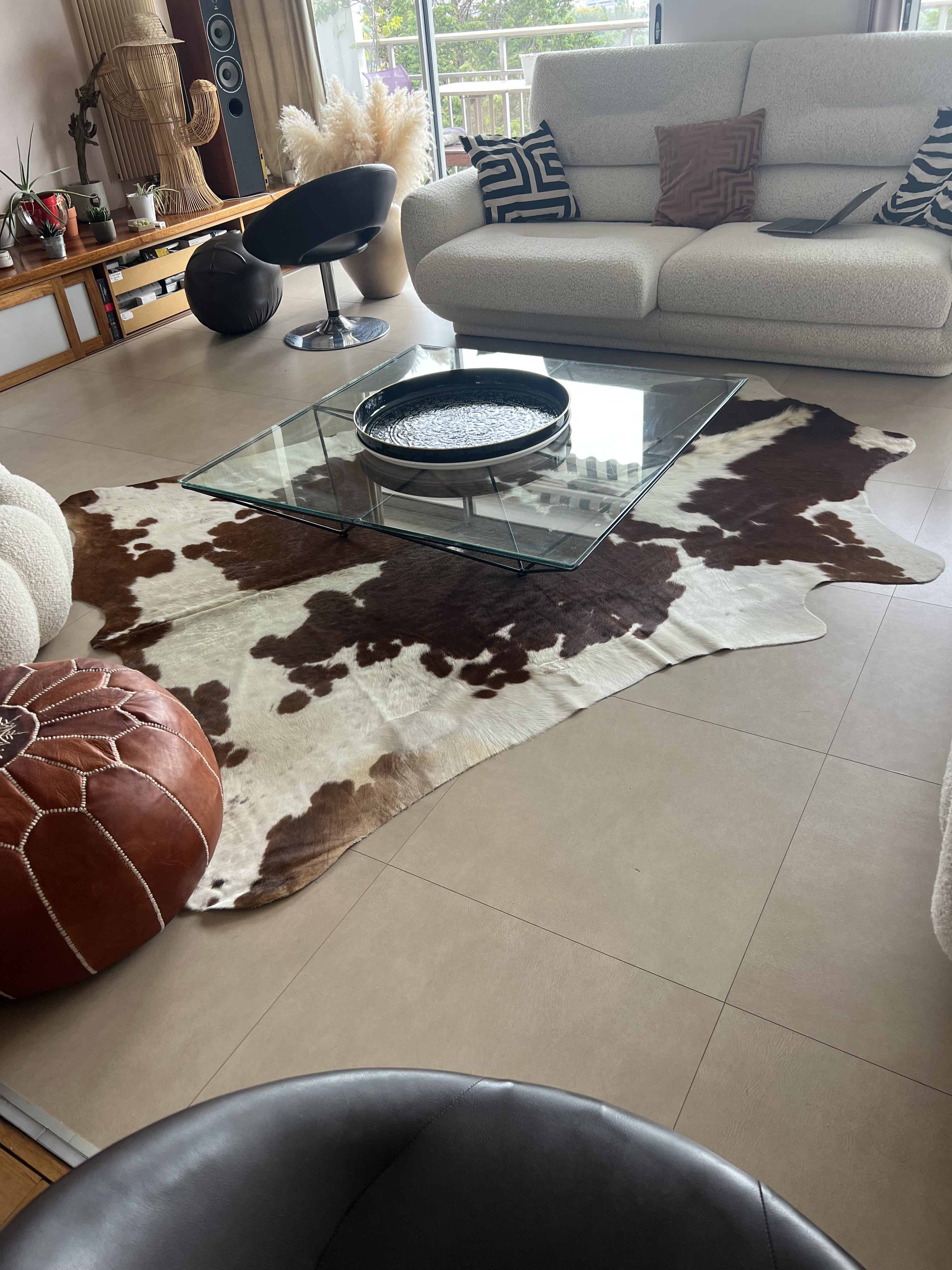 Peau de vache marron et blanche sous une table basse en verre, devant un canapé clair sur un sol carrelé.