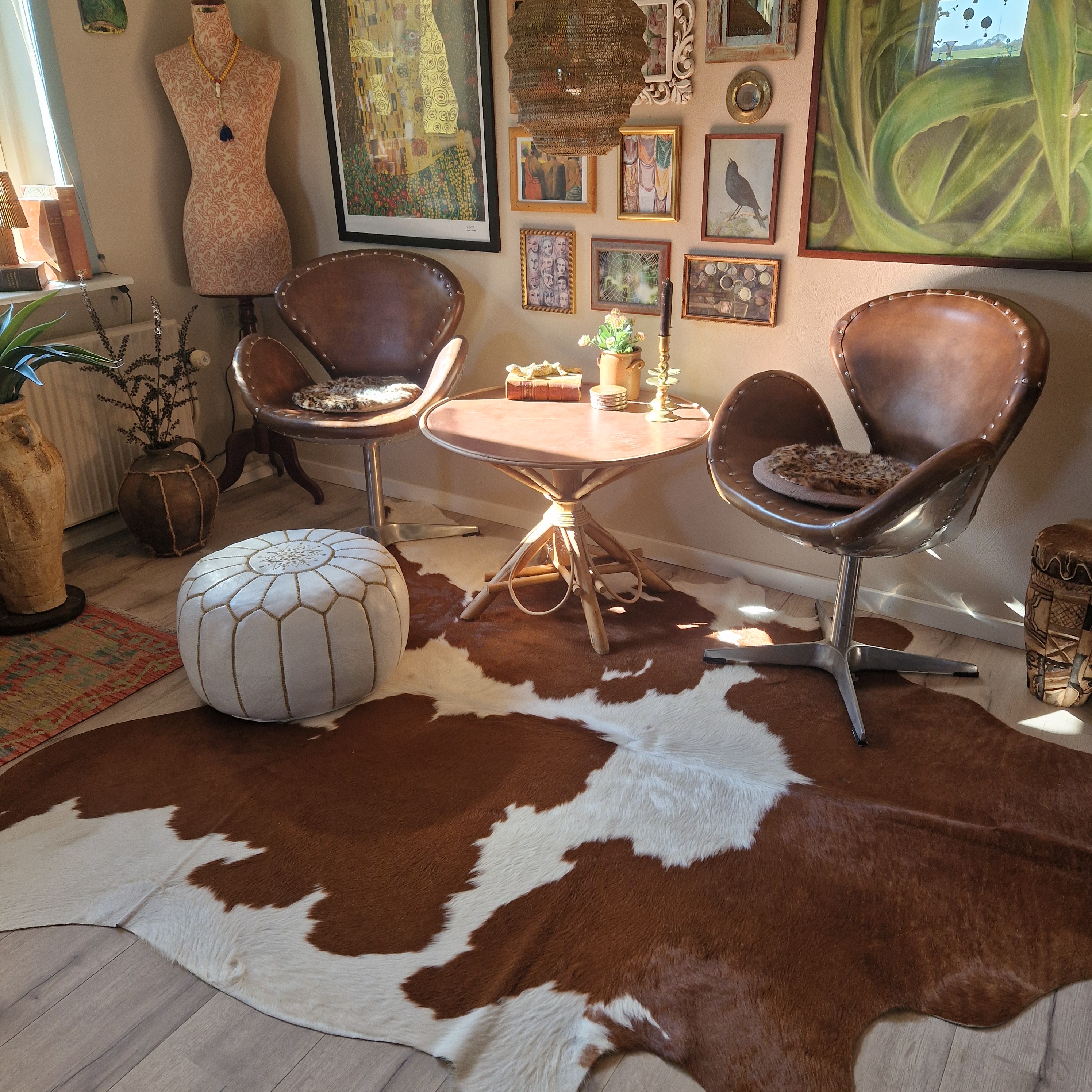 Peau de vache marron et blanche près de deux fauteuils en cuir et d’une table ronde dans un salon décoré de tableaux.