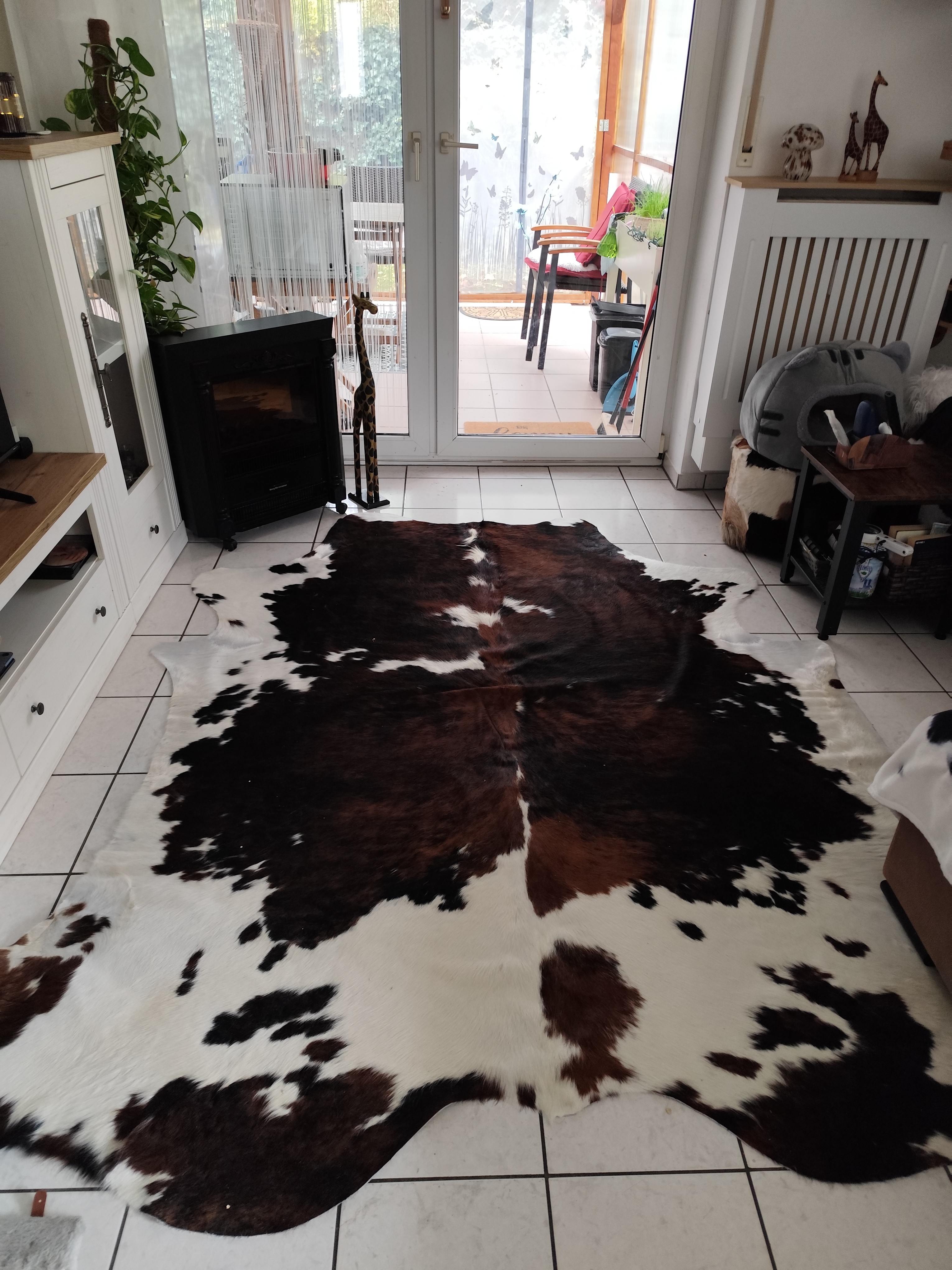 Peau de vache marron et blanche sur un carrelage clair devant une porte vitrée, avec des plantes et des meubles blancs