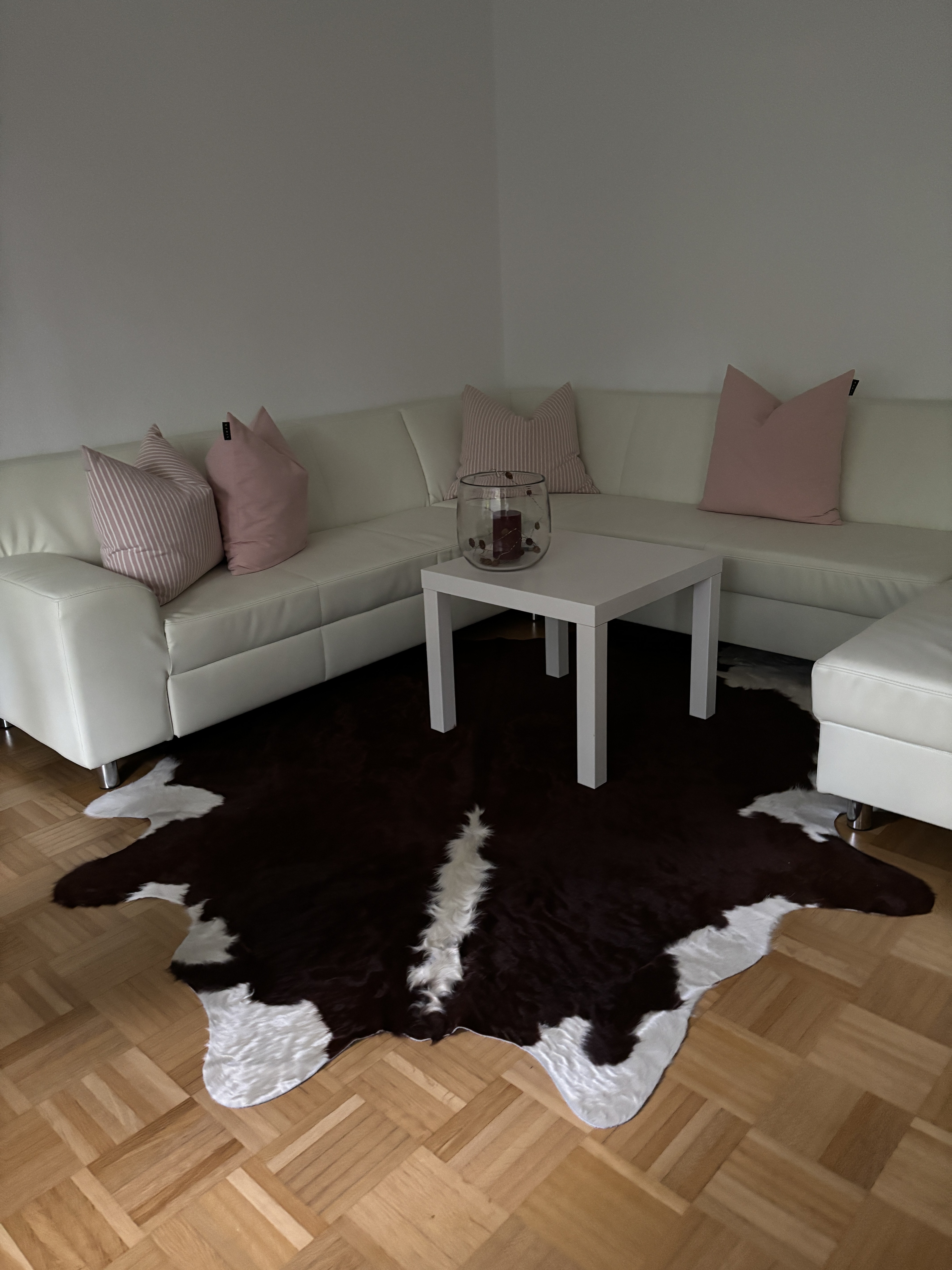 Peau de vache marron et blanche sur un parquet devant un canapé d’angle blanc avec des coussins roses