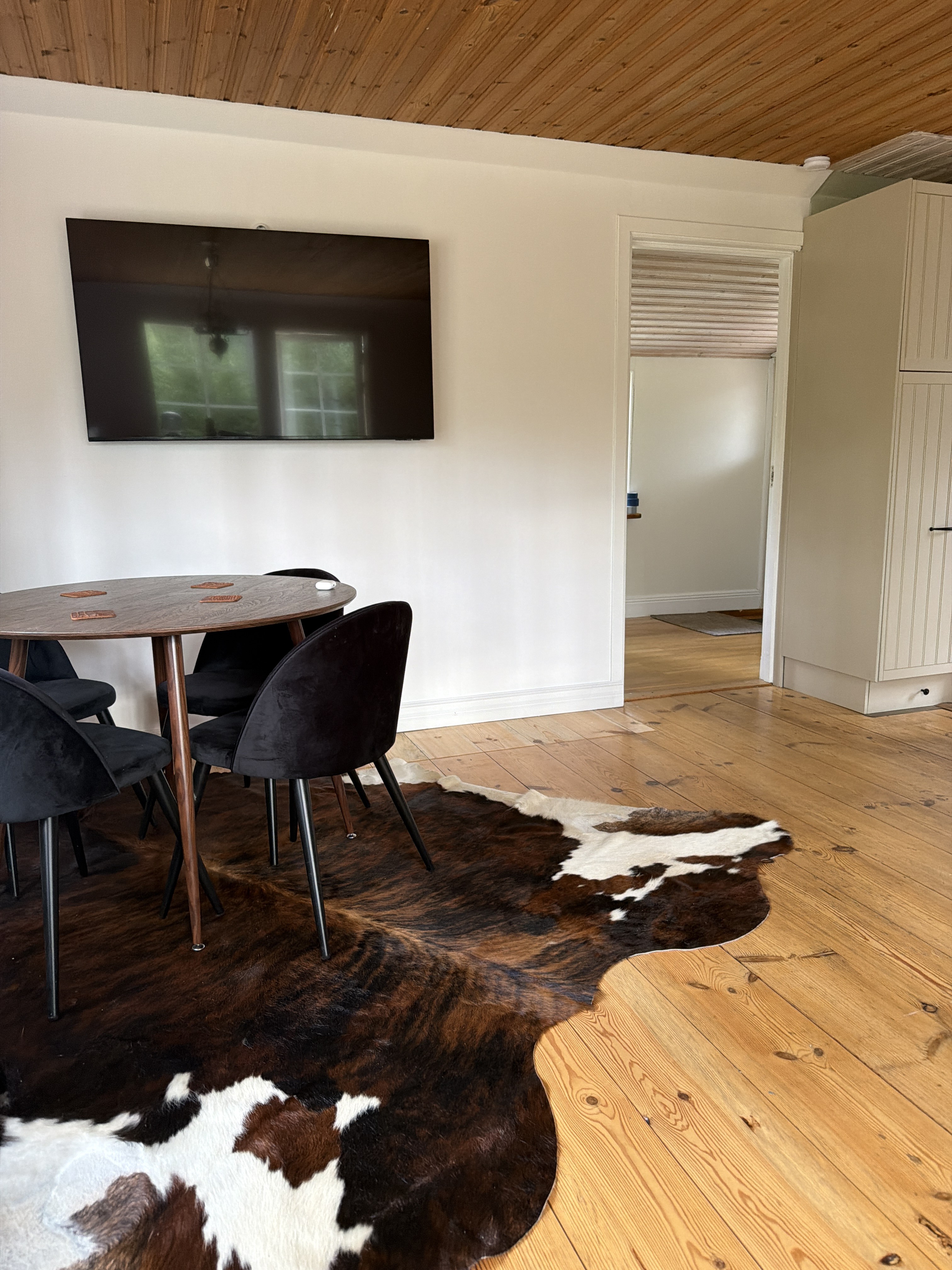 Peau de vache brune et blanche sous une table à manger ronde en bois, avec des chaises noires, dans une pièce lumineuse.