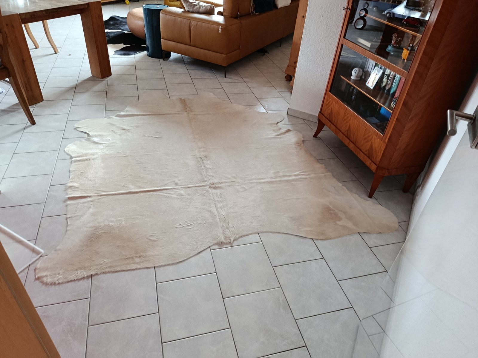 Peau de vache crème sur un carrelage gris clair dans un salon-salle à manger avec table en bois, canapé en cuir cognac et vitrine en bois