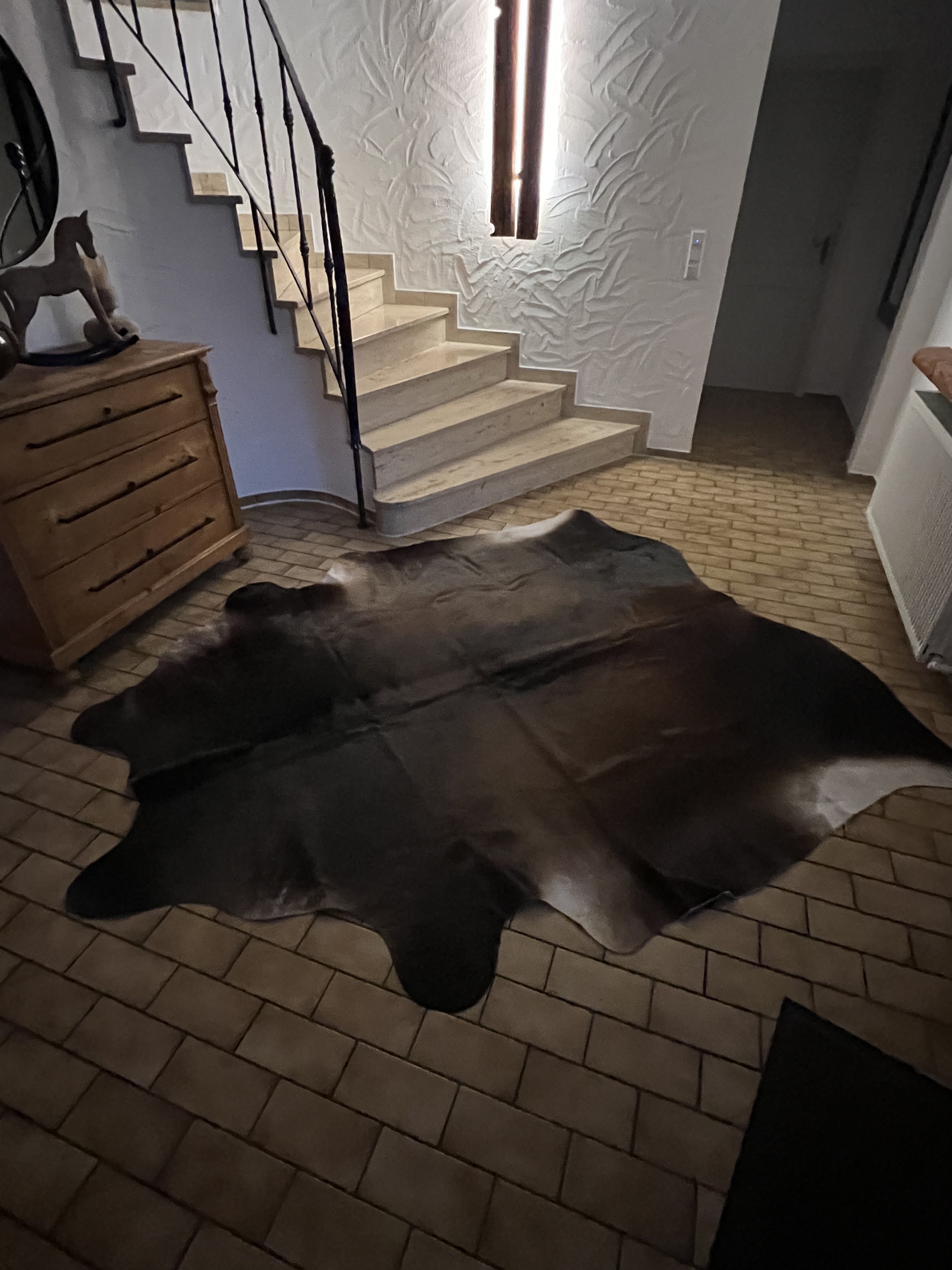 Peau de vache foncée sur un sol en pierre, près d’un escalier en pierre claire et d’une commode en bois
